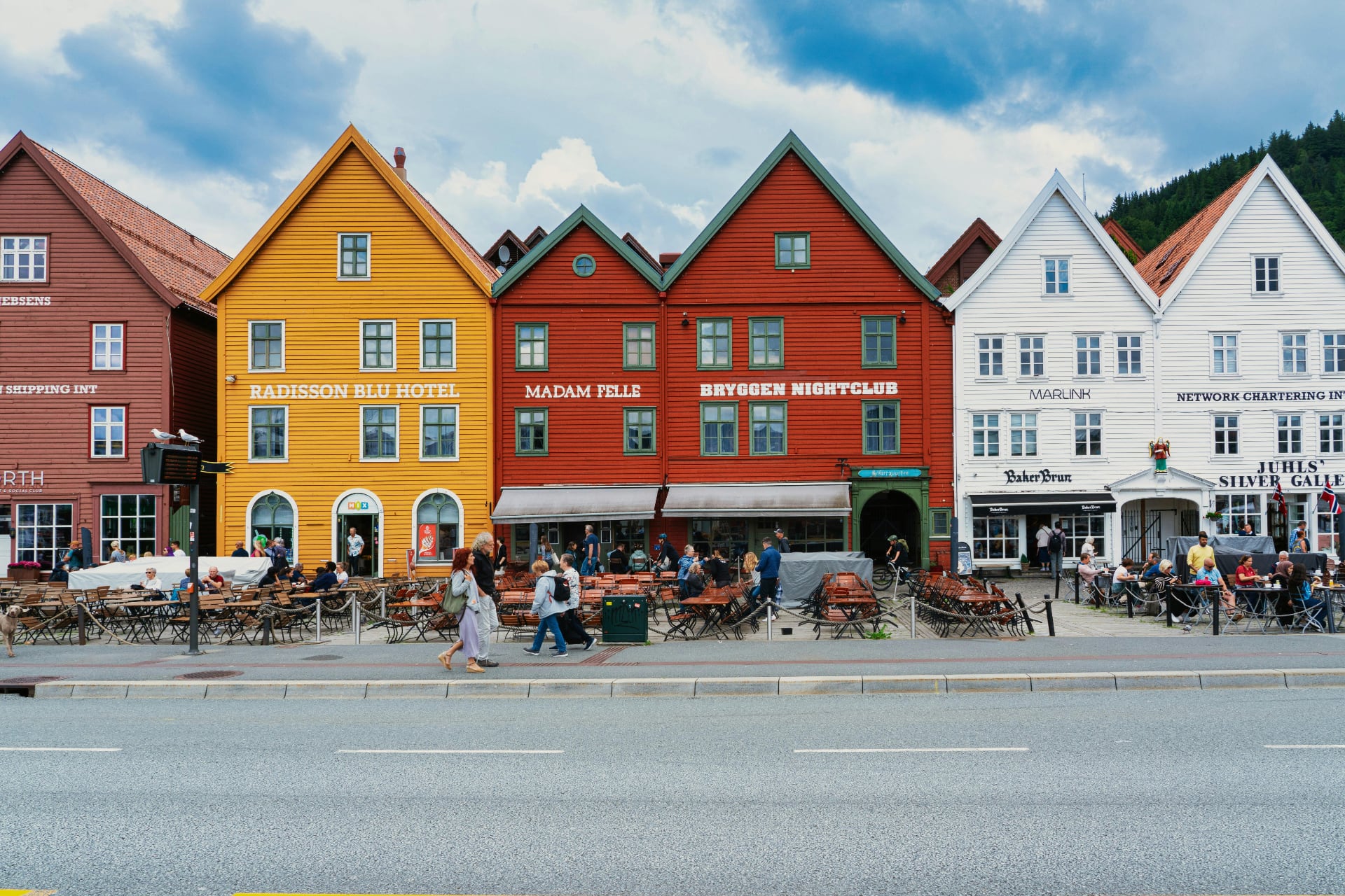 Colourful wooden buildings at Bryggen in Bergen, Norway, with outdoor cafés, shops, and pedestrians along the street on a cloudy day